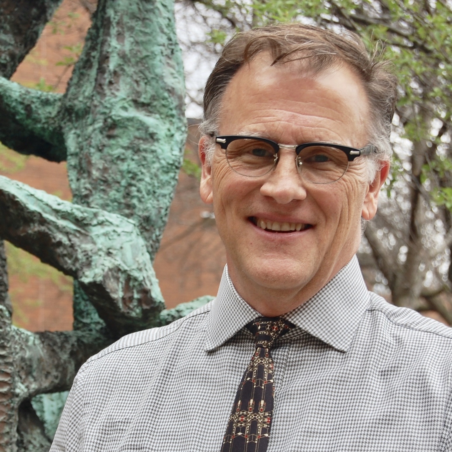 Ray Muno wearing glasses and a button down shirt with tie standing in front of a tree