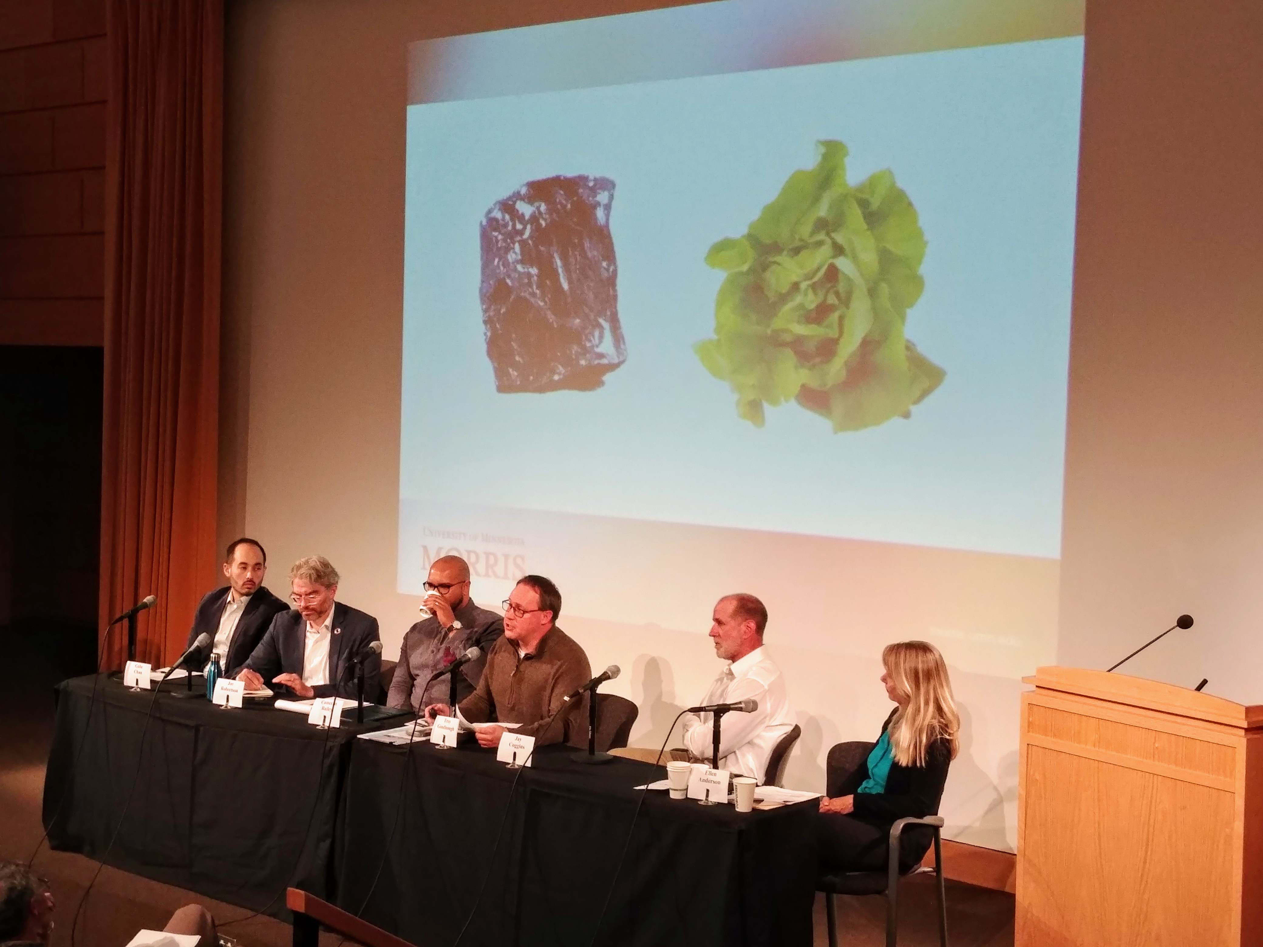 A panel of five members in Cowels Auditorium discusses climate change. There are 5 men (panelists) and one woman (moderator). Behind them is a slide showing, enigmatically, a wad of tin foil and a ball of lettuce.