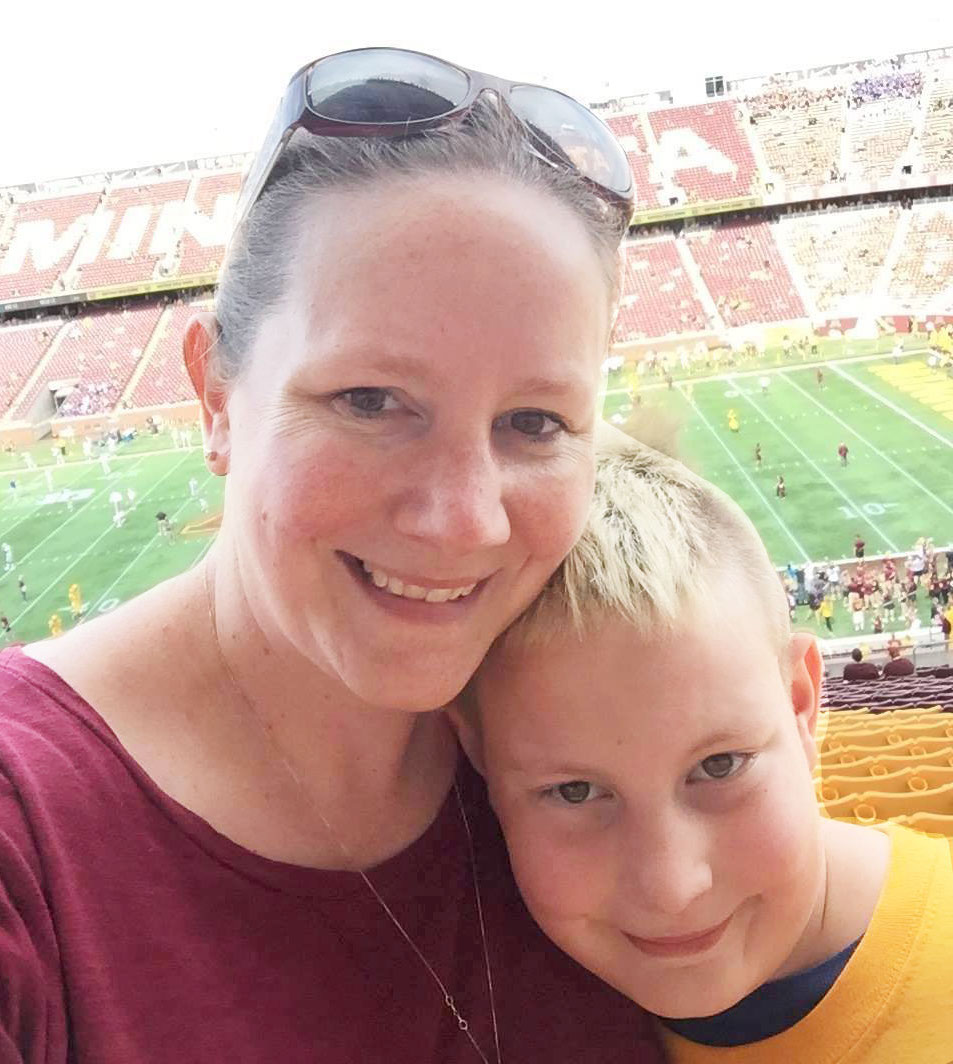 Professor Jennifer Goodnough and her son, a blonde-haired boy roughly age 10, pose at a Gopher sports game. In the background are the football field and stands which spell out \