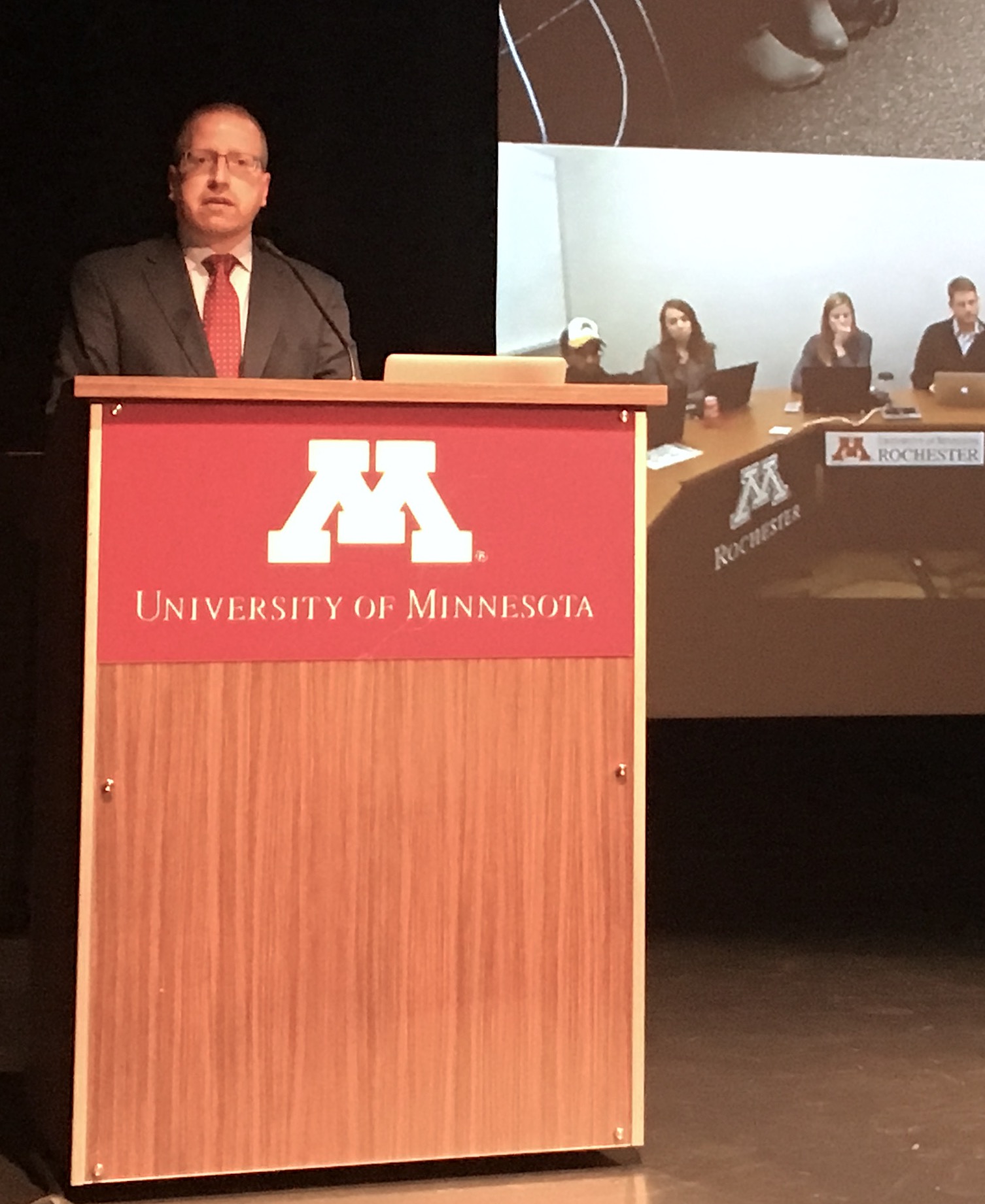 Kevin Dostal Dauer, co-chair, Student Sexual Misconduct Subcommittee, addresses the University Senate in Coffman Theater. In the background, the Rochester campus, connected by ITV, is visible.