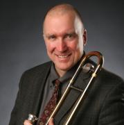 Dean Sorenson wearing a dark gray suit and short with a maroon patterned tie, holding a trombone