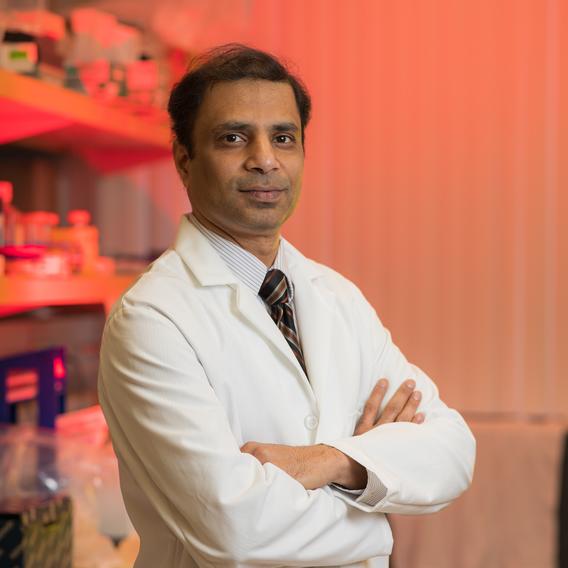 Photograph of Subree Subramanian in a white lab coat with a white shirt and tie. He is standing with his arms crossed in an orange-tinted room; there are pharmaceuticals on the shelf behind him