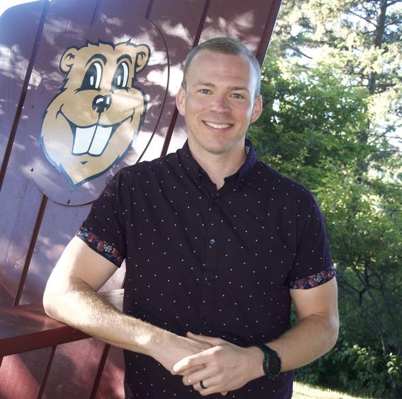 Tom Hegblom poses outdoors near a large, maroon adirondack chair with Goldy Gopher's face on it