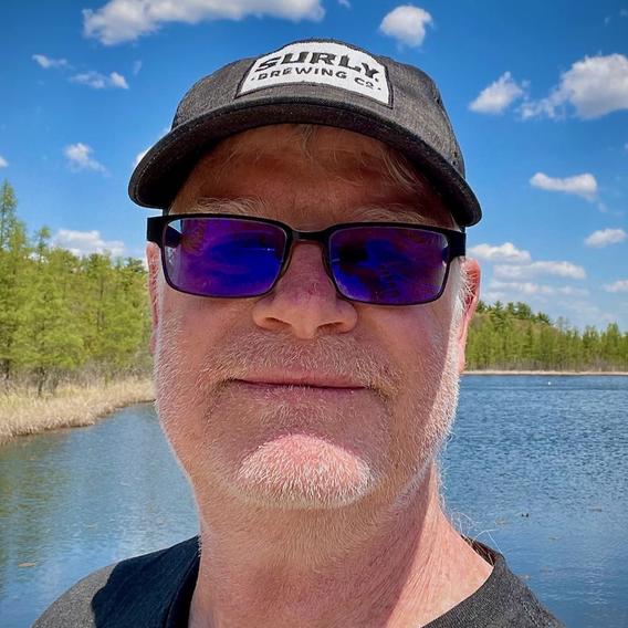 Professor Jim Cotner in a hat and sunglasses, in front of a tree-lined lake. The sky is brilliant blue and dotted with wispy clouds.