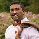 Fiyyaz Karim poses in a field of wildflowers, wearing a bowtie and holding his jacket over his shoulder