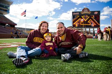 A mother, father, and baby wearing full Gopher gear sit on the football field.
