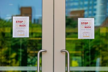 A sunny day on campus is reflected in the glass in a set of double doors. On the doors, two signs remind people to put masks on before entering.