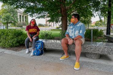 A woman in a U of M tshirt and a mask sits on a bench outdoors, 6 feet away from a man who is also wearing a mask.