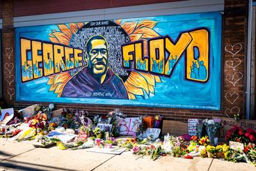A mural reading "George "Floyd" with a poirtrait of him between the words. There are many flowers, signs, and tributes at the foot of the mural. Photo by munshots on Unsplash.