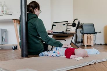 A woman site on the floor working on a laptop on and adjustable-hight desk. She rests her hand on the back of an infant who is sleeping face down on the floor next to her.