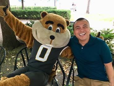 Adolfo Carrillo Cabello poses with Goldy Gopher. They are seated in chairs on Northrop Mall. It is sunny, they have their arms around one another's shoulders, Adolfo is smiling, Goldy is giving the thumbs up.